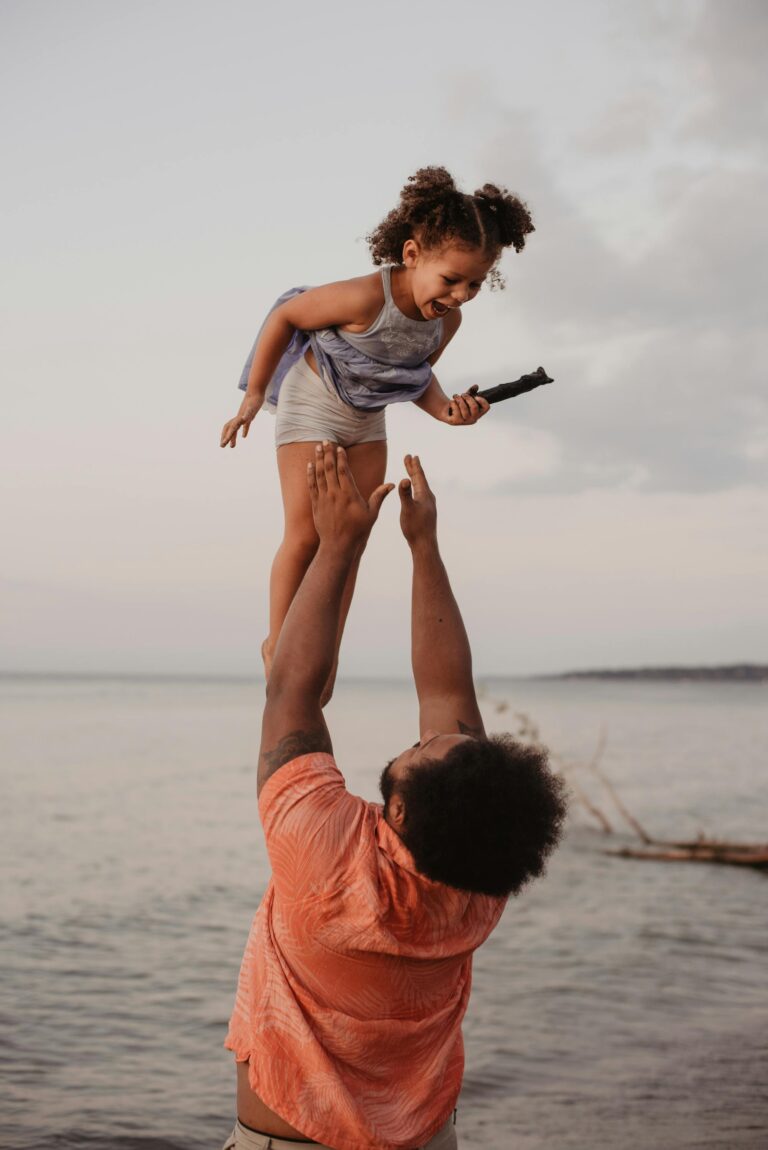 A joyful father lifts his daughter in the air by the beach during sunset, capturing a moment of love and happiness.